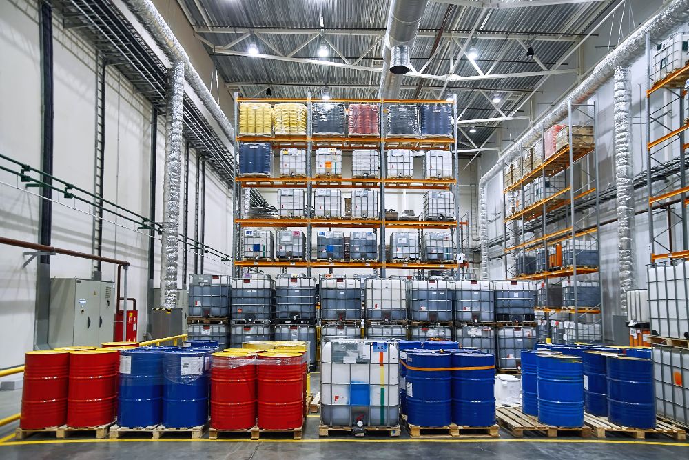 Industrial warehouse with metal racking and filled with sealed barrels and containers, mainly in red, blue, yellow, and gray colors.