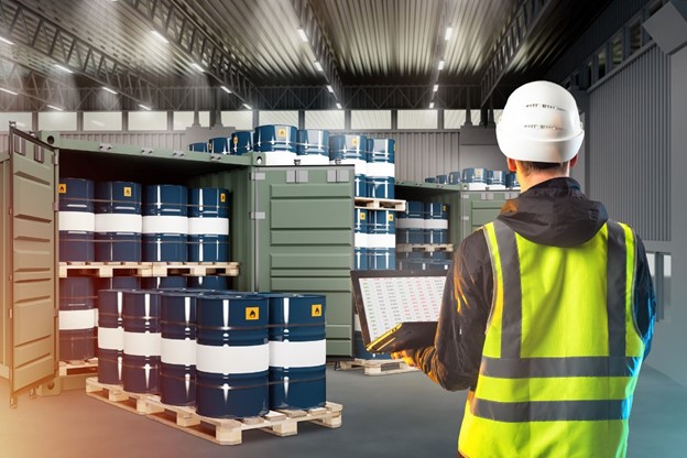 A worker in a high-visibility vest and hard hat uses a laptop while inspecting chemical storage area filled with barrels inside a well-lit warehouse.