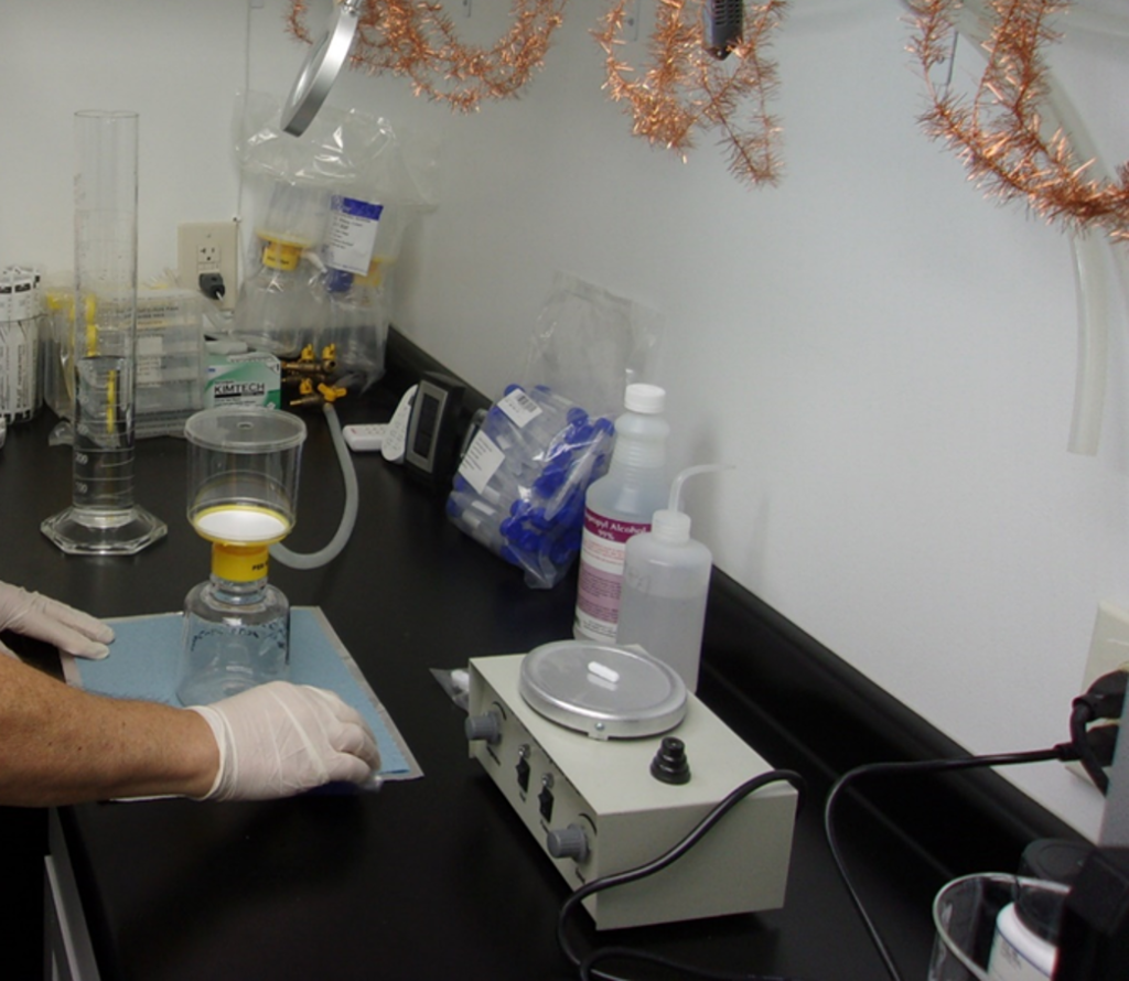 A person wearing gloves operates microbiology lab equipment including a beaker, a filtration apparatus, and a heating plate on a laboratory bench with various supplies.